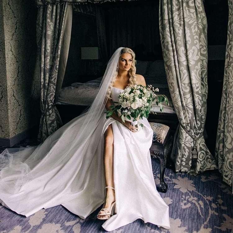 A bridge sits on a stool at the end of a bed, she is wearing a white wedding dress in the bridal suite at Clontarf Castle Wedding Venue in Dublin