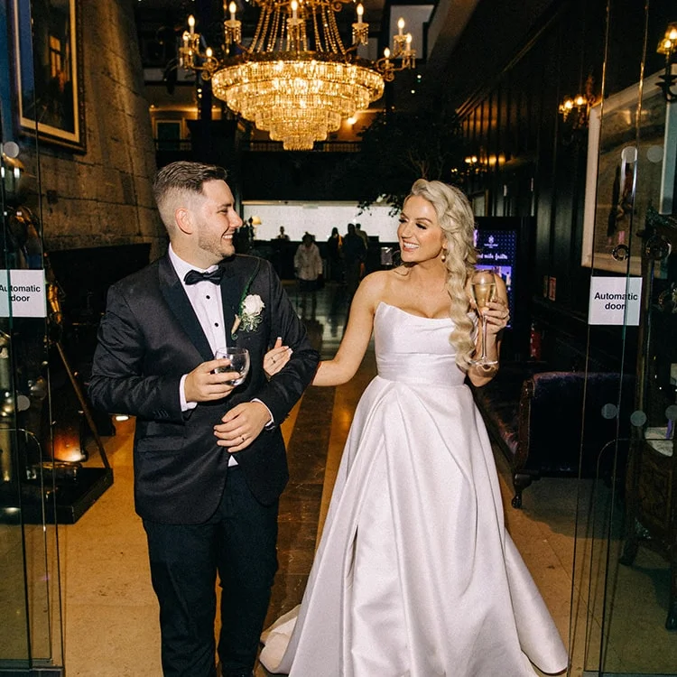 A husband and Wife link arms while holding a drink and walking through the doors of Clontarf Castle Wedding Venue in Dublin