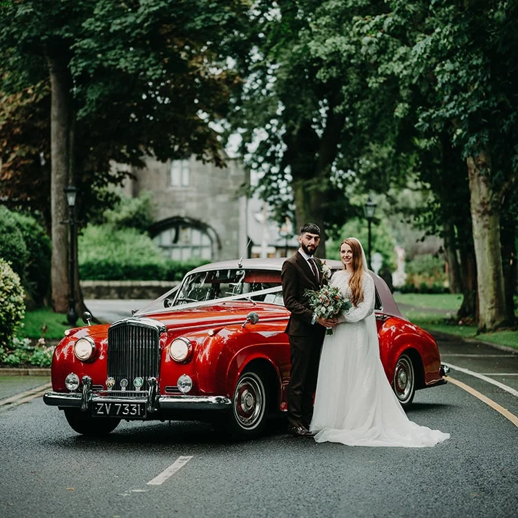 A bride and groom stand infront of a red car on the driveway of Clontarf Castle Wedding Venue in Dublin