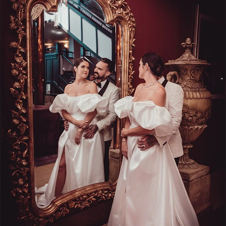 A couple in white wedding attire stand infront of a mirror in Clontarf Castle Wedding Venue