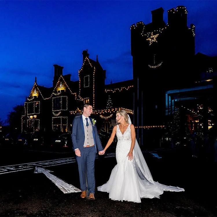 A bride and Groom hold hands infront of the Castle illuminated with Christmas Decorations at Clontarf Castle Wedding Venue in Dublin.