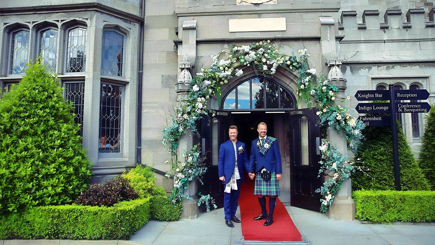 A man in a suit and another in a kilt stand hand in hand at the exterior of Clontarf Castle, the stand on a red carpet as they enter into a civil ceremony