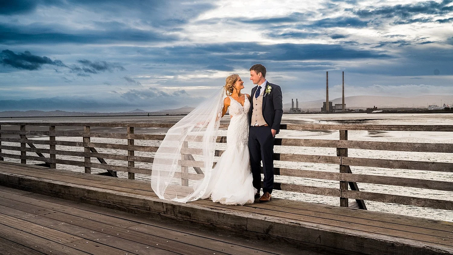 A bride and groom gaze into each others eyes as they stand on the wooden bridge to Bull island, with the red and white poolbeg chimneys in the background